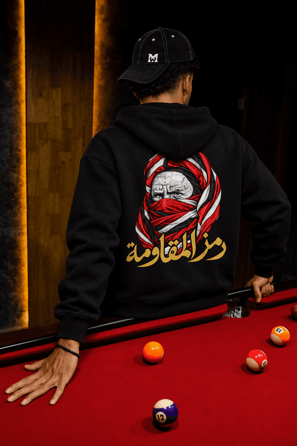 Homme de dos appuyé contre un billard rouge, portant le hoodie noir au portrait rouge et blanc et à la calligraphie dorée.