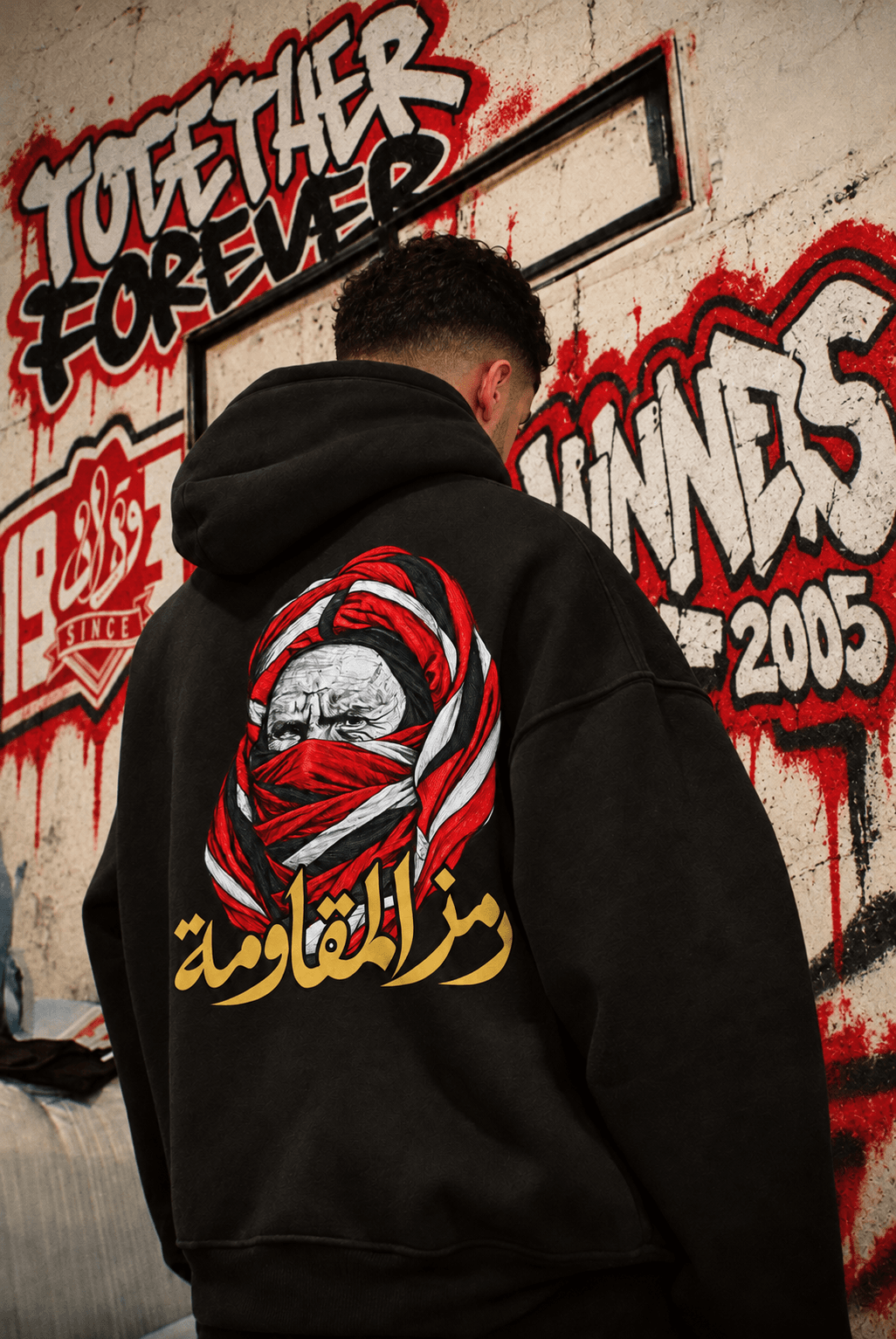 A supporter seen from behind in the black hoodie, with red, white and gold artwork, beside a wall painted with TOGETHER FOREVER and WINNERS 2005.