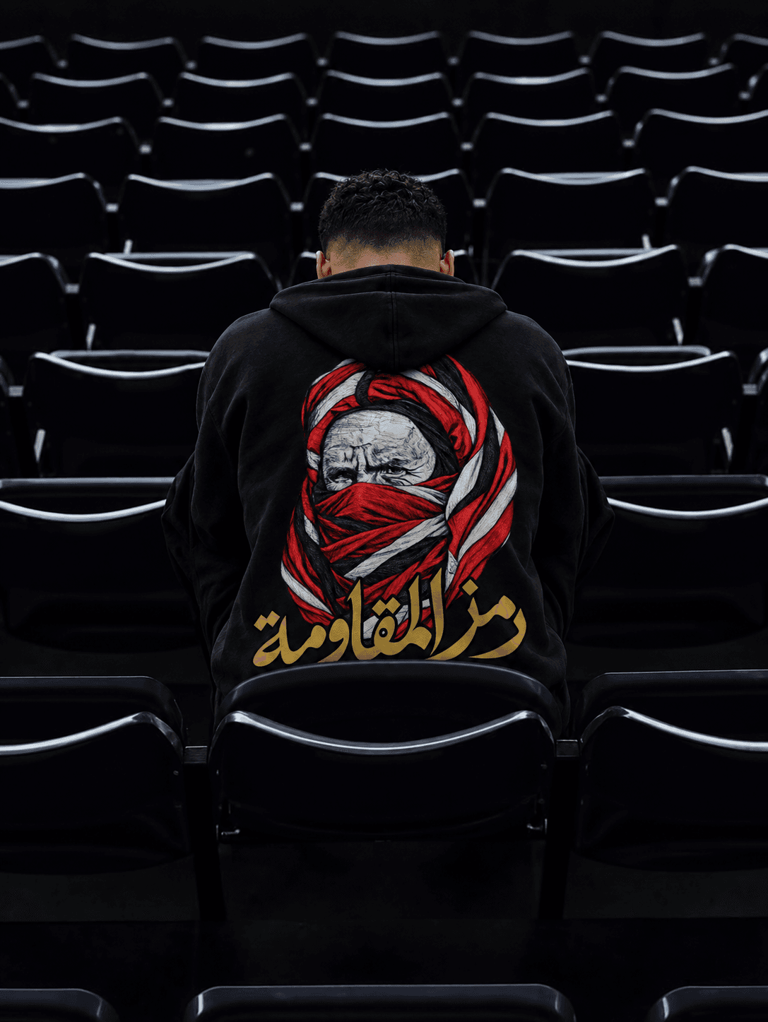 Homme de dos assis parmi des rangées de sièges noirs, le portrait rouge et blanc et les lettres dorées du hoodie visibles au centre.